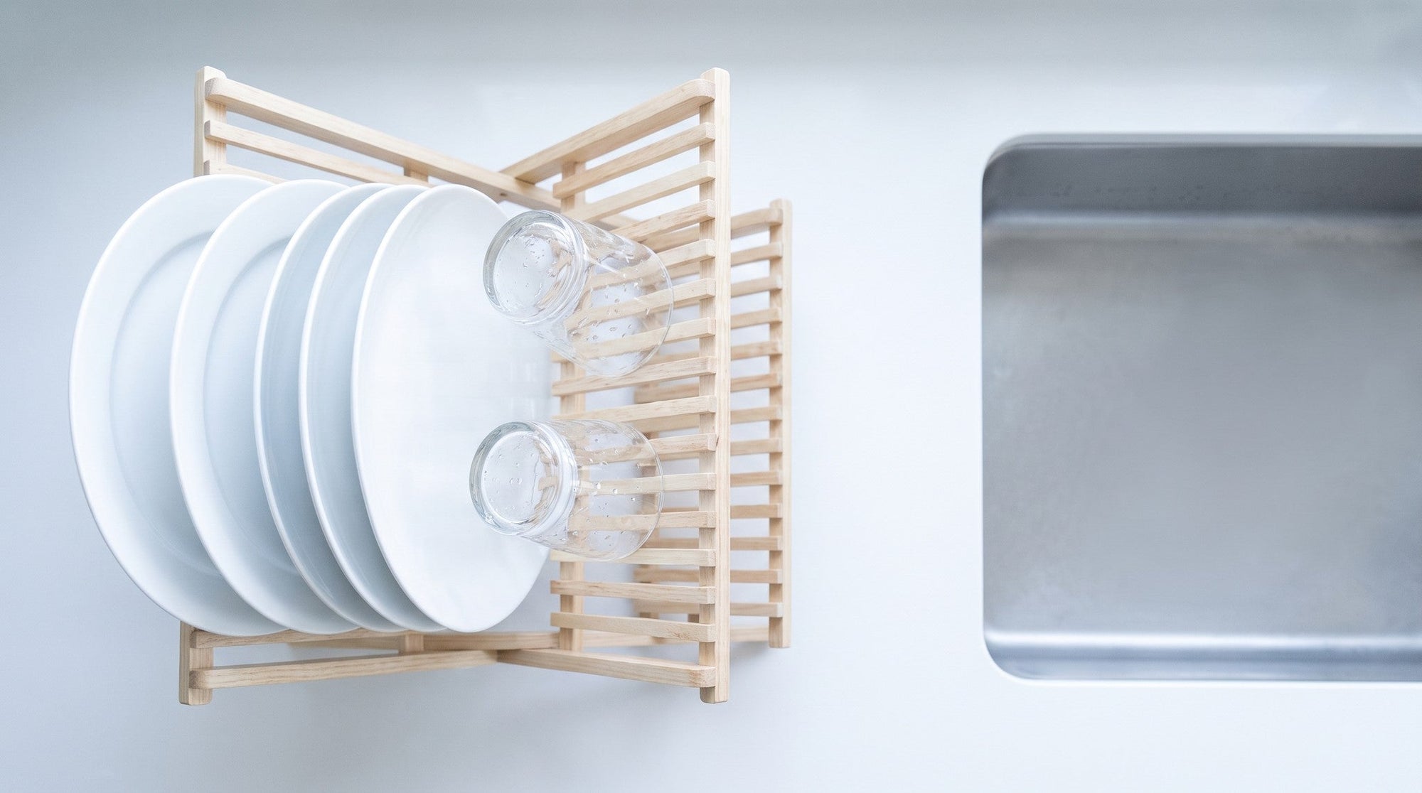 Clean white plates and glasses draining in a wooden dish rack beside an empty, dry kitchen sink, seen from above in bright daylight.
