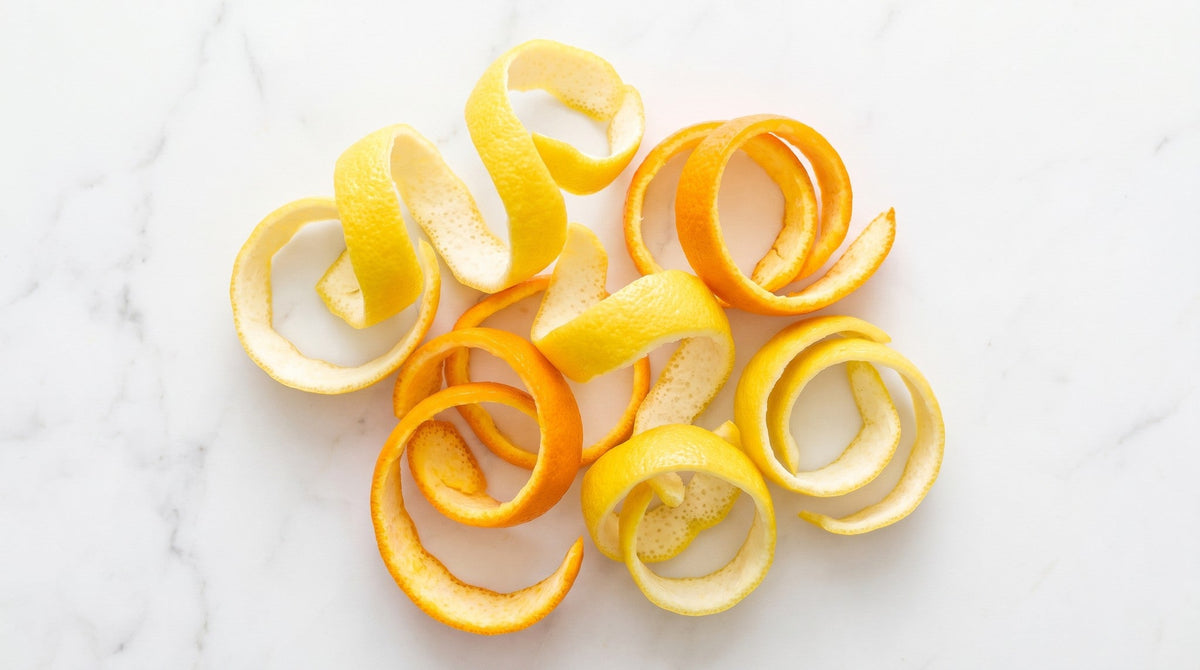 Curled ribbons of lemon and orange peel scattered across a white marble surface, seen from above in bright daylight.