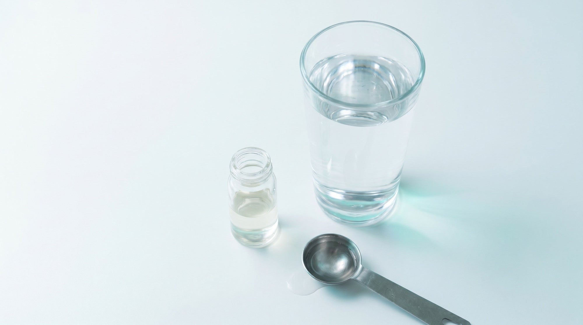 Overhead view of a small concentrate vial beside a tall glass of water and a measuring spoon on a clean white surface.