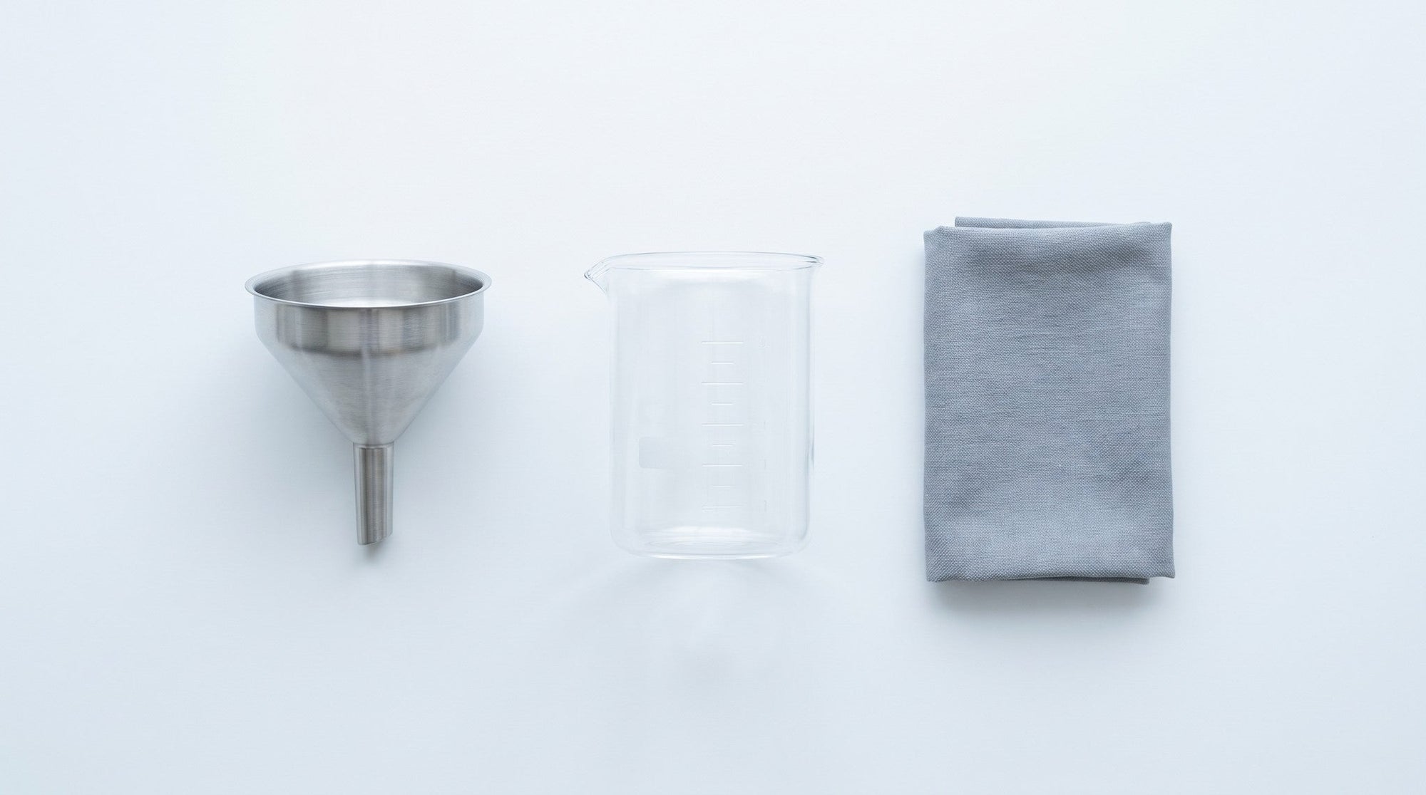 A stainless funnel, a plain glass beaker and a folded grey cloth laid out on a white surface, seen from above.