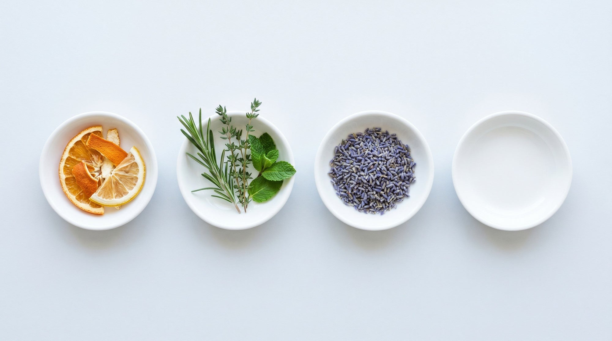 Overhead view of small white dishes holding citrus peel, fresh herbs and dried lavender beside one empty dish.