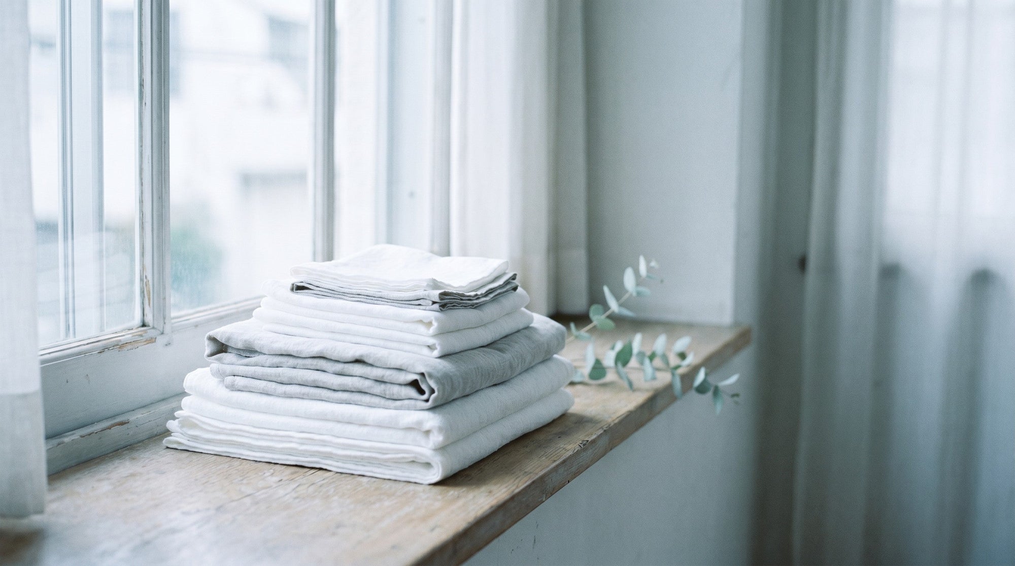 A soft stack of folded white and pale-grey linens near a window in gentle diffused light.
